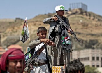 TOPSHOT - A Yemeni boy lines up a round of ammunition atop the barrel of a Kalashnikov assault rifle, with a flag sticking from his jacket showing a picture of the Huthi rebel leader Abdulmalik al-Huthi, while another buy stands with Kalashnikov over a crate of juice boxes during a tribal meeting in the Huthi rebel-held capital Sanaa on September 21, 2019, as tribesmen donate rations and funds to fighters loyal to the Huthis along the fronts. (Photo by MOHAMMED HUWAIS / AFP)        (Photo credit should read MOHAMMED HUWAIS/AFP via Getty Images)