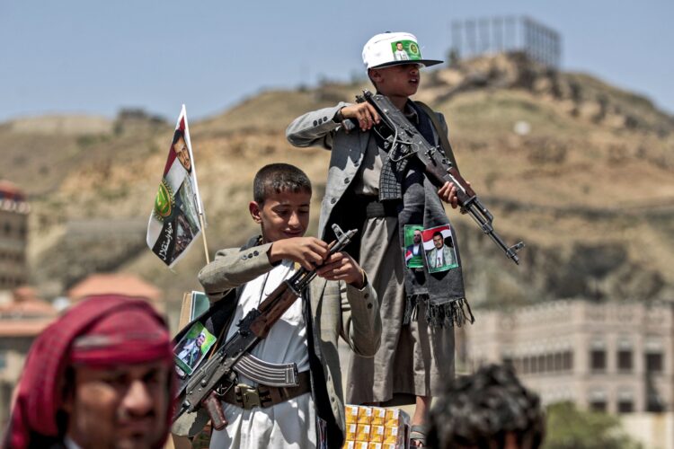 TOPSHOT - A Yemeni boy lines up a round of ammunition atop the barrel of a Kalashnikov assault rifle, with a flag sticking from his jacket showing a picture of the Huthi rebel leader Abdulmalik al-Huthi, while another buy stands with Kalashnikov over a crate of juice boxes during a tribal meeting in the Huthi rebel-held capital Sanaa on September 21, 2019, as tribesmen donate rations and funds to fighters loyal to the Huthis along the fronts. (Photo by MOHAMMED HUWAIS / AFP)        (Photo credit should read MOHAMMED HUWAIS/AFP via Getty Images)